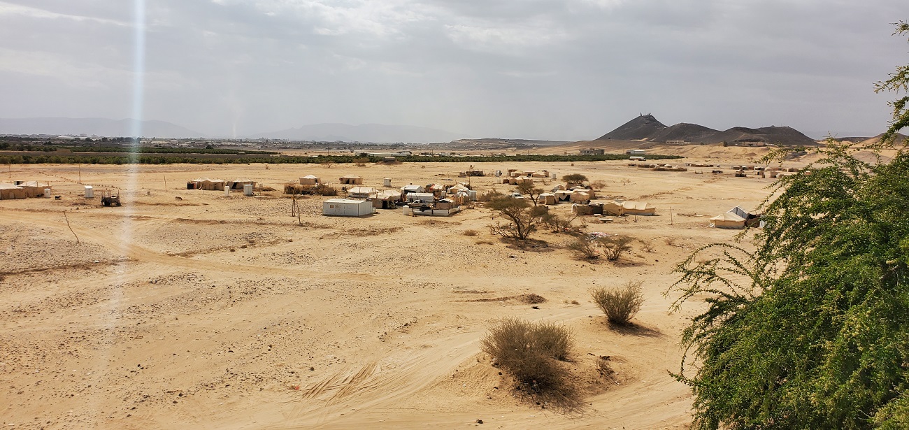 A desert landscape with several tents and sparse trees under a clear blue sky.