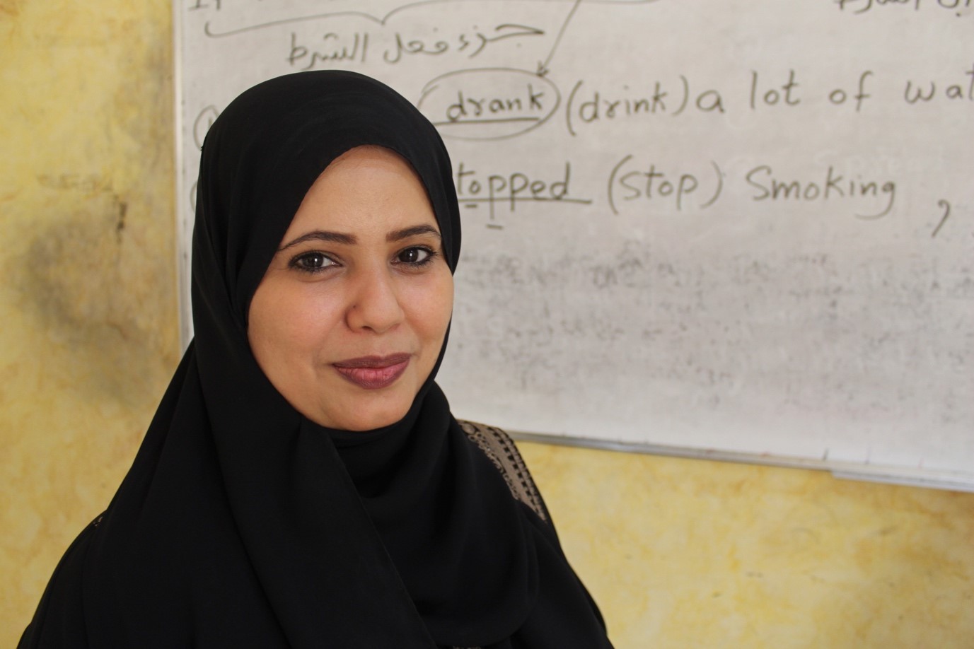 A woman wearing a black hijab stands confidently in front of a whiteboard, ready to present or teach.