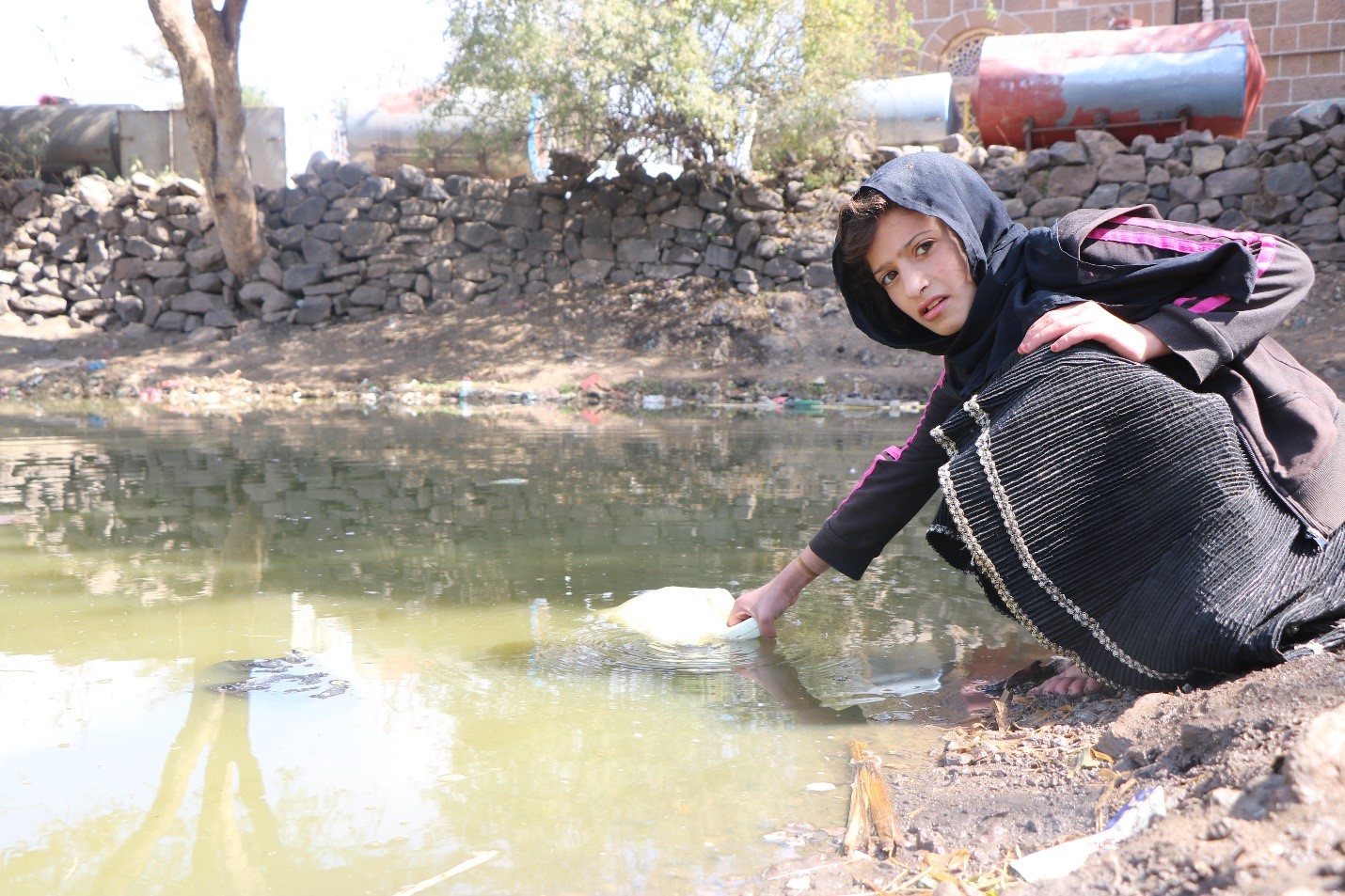 A girl in a black hoodie kneels down by a stream, collecting water in a container.