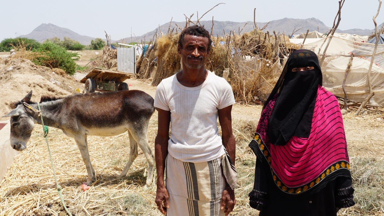 A man and woman standing in front of a donkey in a rural setting.