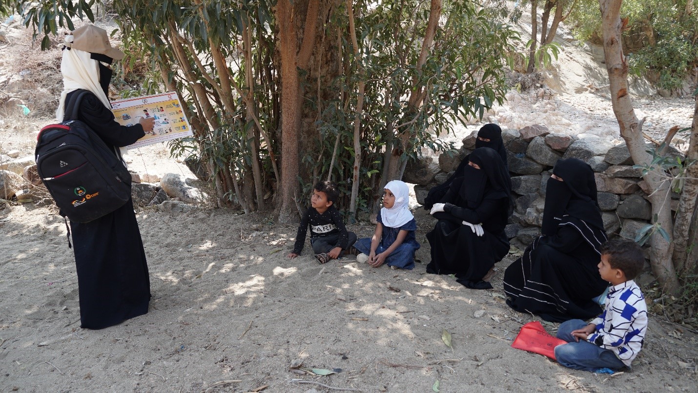 A group kids sitting under a tree listening to a woman standing in front of them