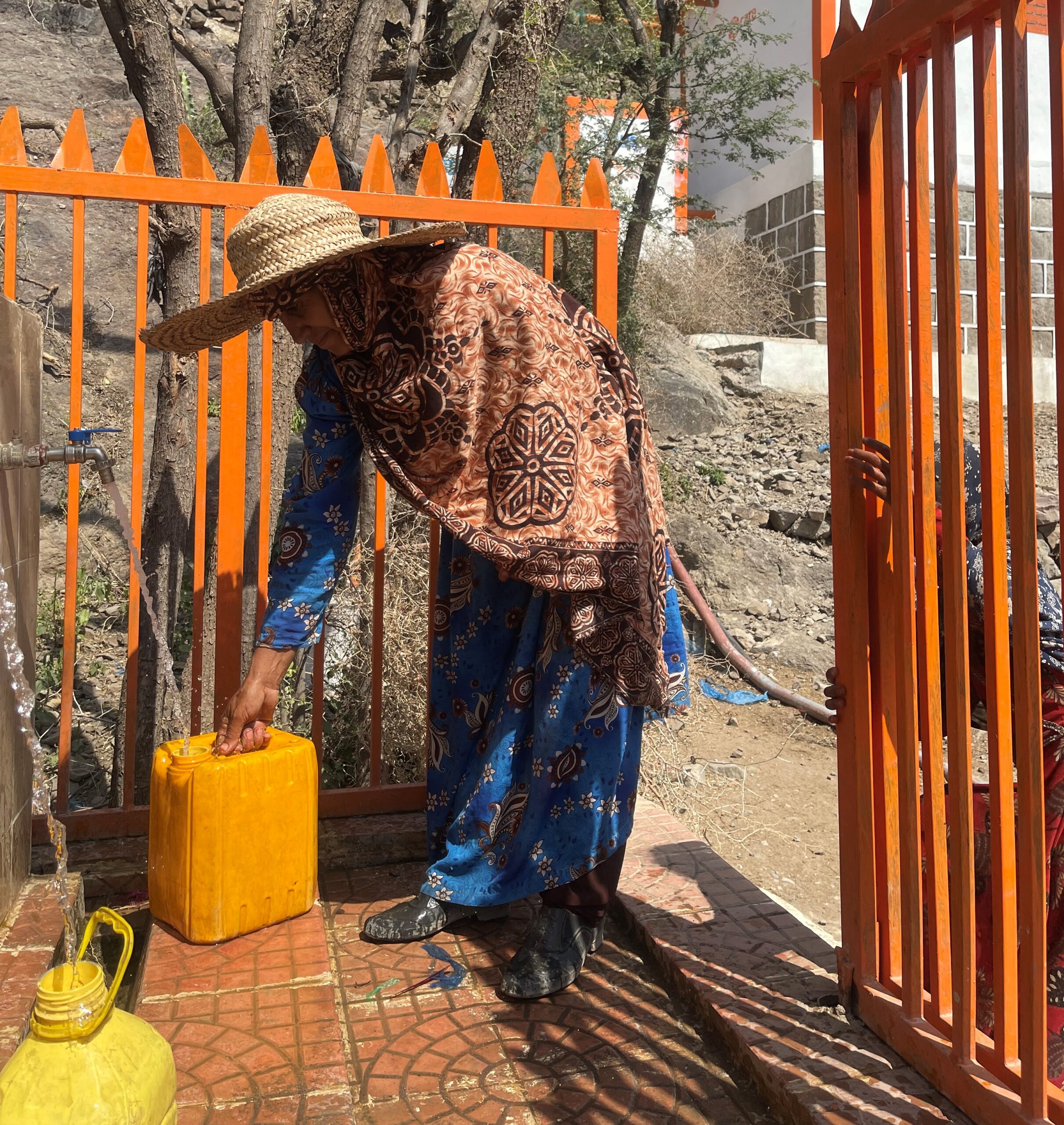 a woman wearing a hat and holding a yellow container fetching water from a tap