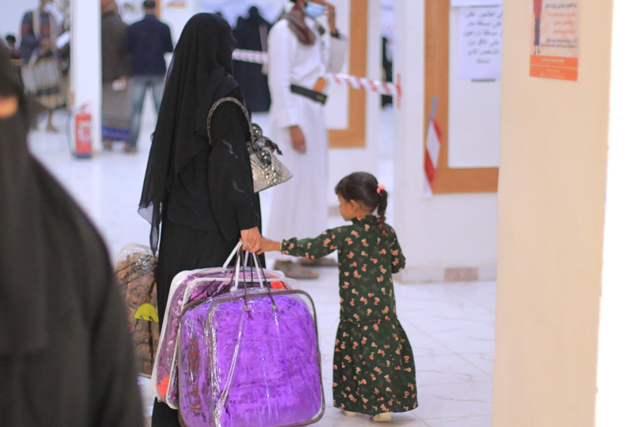 A woman and a child walking through a hallway, holding hands and luggage