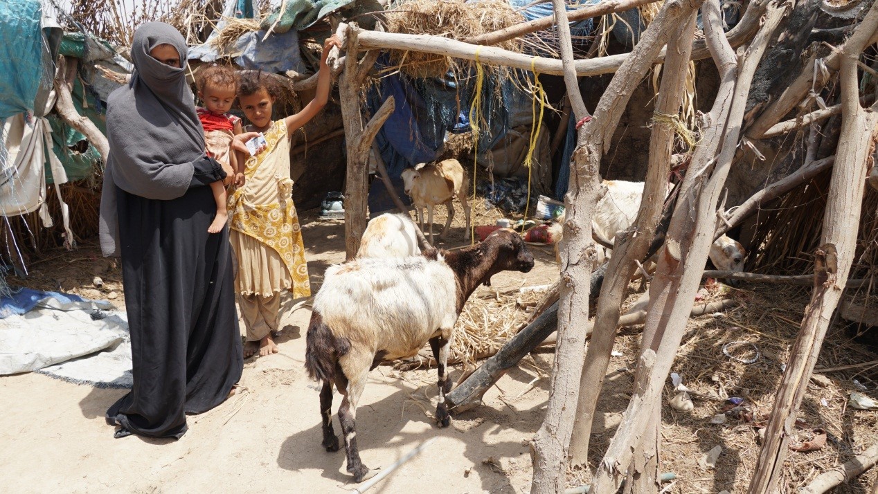 A woman holding a baby and a small girl standing beside her next to a goat shed