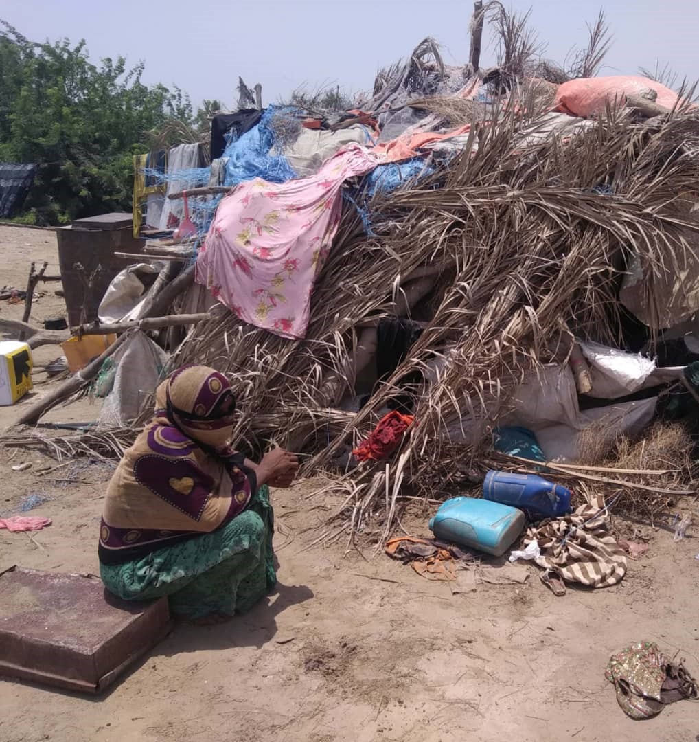A woman sitting in front of a hut surrounded by various items.
