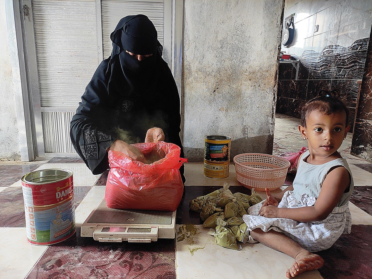 A woman in a black head covering measuring something on a scale and a baby sitting next to the scale