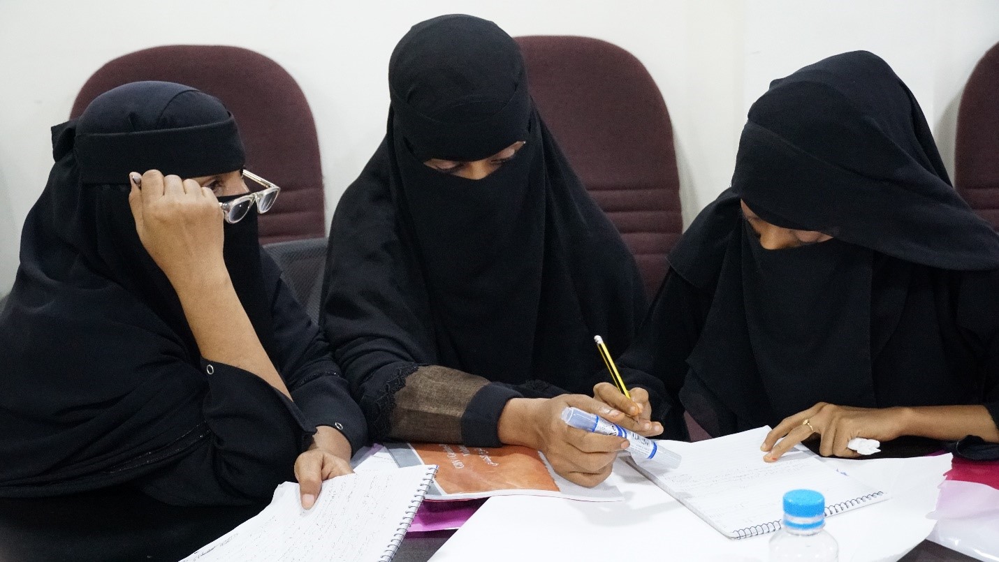 Three women in burqas sitting at a table, reviewing papers and engaging in a discussion.