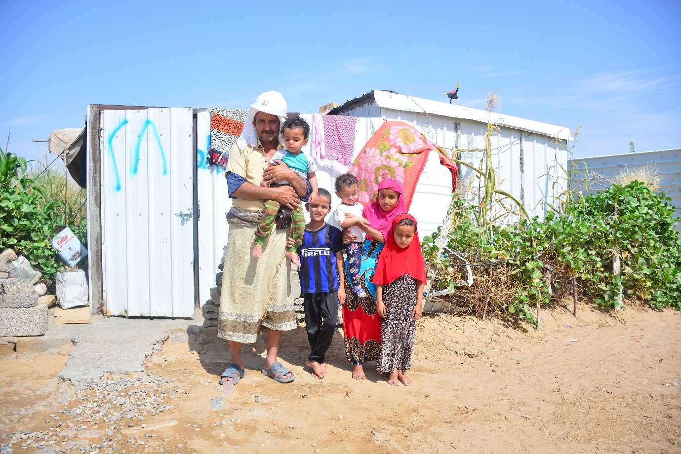 A man holding a child and standing in front of a white shed with other kids standing beside him