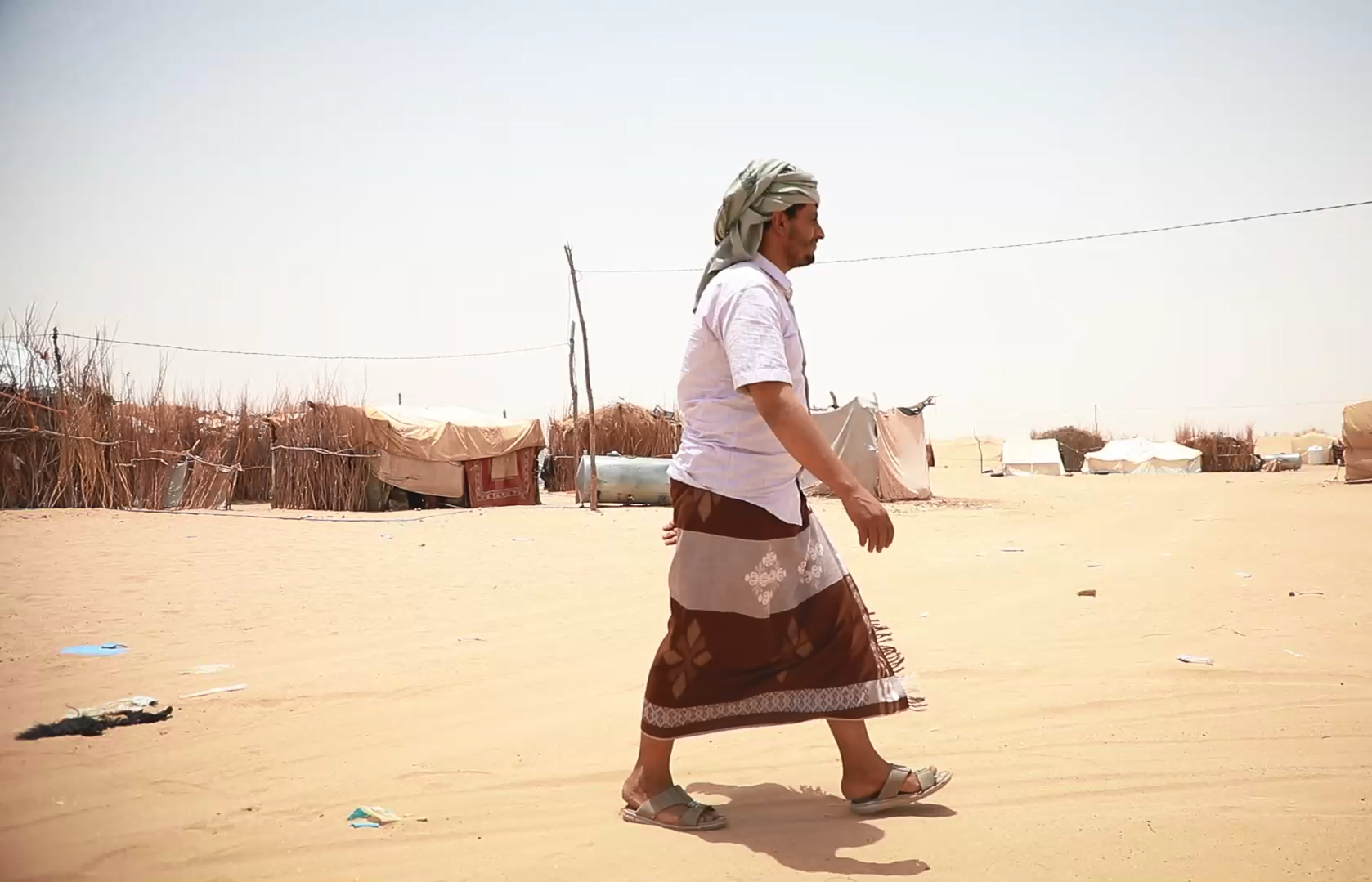 A man walking in the desert near a tent, under the scorching sun.